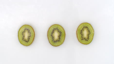 Slow-motion-water-splash-on-three-slices-of-green-kiwi-lying-on-a-white-background-in-the-water